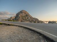 a motorcycle parked by the side of a mountain road at sunset, with a road and rocks in the background