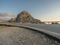 a motorcycle parked by the side of a mountain road at sunset, with a road and rocks in the background