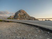 a motorcycle parked by the side of a mountain road at sunset, with a road and rocks in the background