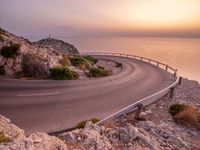 the road is curving near the edge of the hill at sunset with sea in the background