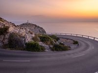 the road is curving near the edge of the hill at sunset with sea in the background