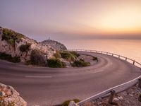 the road is curving near the edge of the hill at sunset with sea in the background