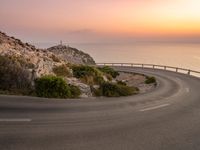 the road is curving near the edge of the hill at sunset with sea in the background