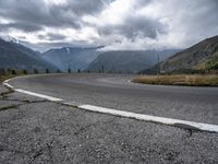 an empty winding road leading into the mountains and mountain range in a cloudy day time