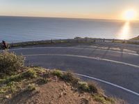 a cyclist riding down a curved road by the ocean at sunset over the water and hills