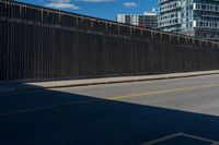 a man is riding his bike on the side of a road beside tall buildings and a fence