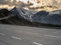 the person is riding their bike on the road in the mountains under cloudy skies and snow covered hills