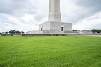 a monument stands surrounded by green grass with people in it and clouds overhead over a monument