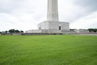 a monument stands surrounded by green grass with people in it and clouds overhead over a monument