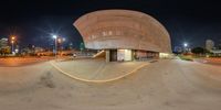 a large circular building in the middle of city street with lights at night time and parking lot