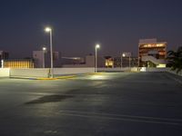 an empty parking lot is lit up at night with bright street lights overhead them are buildings and palm trees