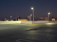 an empty parking lot is lit up at night with bright street lights overhead them are buildings and palm trees