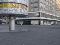 a clock with numbers is sitting on the middle of the street near some tall buildings