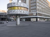 a clock with numbers is sitting on the middle of the street near some tall buildings