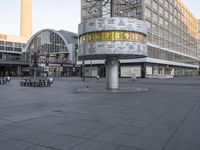 a clock with numbers is sitting on the middle of the street near some tall buildings