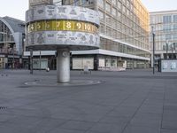 a clock with numbers is sitting on the middle of the street near some tall buildings