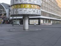 a clock with numbers is sitting on the middle of the street near some tall buildings