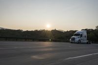 a semi truck traveling down a road next to the sun setting behind the trees that line the highway