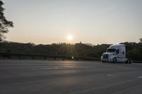 a semi truck traveling down a road next to the sun setting behind the trees that line the highway