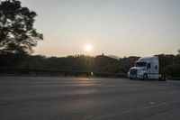 a semi truck traveling down a road next to the sun setting behind the trees that line the highway