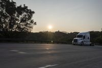 a semi truck traveling down a road next to the sun setting behind the trees that line the highway