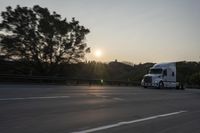 a semi truck traveling down a road next to the sun setting behind the trees that line the highway