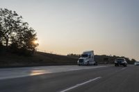 a semi truck traveling down a road next to the sun setting behind the trees that line the highway