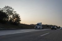 a semi truck traveling down a road next to the sun setting behind the trees that line the highway