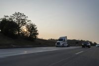 a semi truck traveling down a road next to the sun setting behind the trees that line the highway