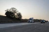 a semi truck traveling down a road next to the sun setting behind the trees that line the highway