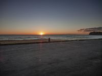 the man is walking on the edge of a road near the beach as the sun sets