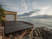 an open wooden deck overlooking the water in front of a beach cottage and garage building