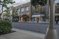 a long sidewalk with trees and bushes on it next to stores on both sides of the street