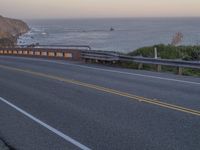 a person riding a motorcycle down the side of the road by the ocean shore at dusk