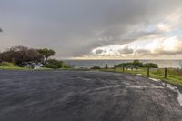 an empty road next to the ocean on a cloudy day at dusk with a bench out side