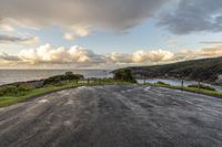 an empty road overlooks the ocean below a hillside with grass on either side and a few clouds in the sky