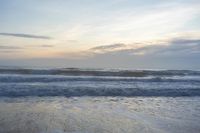 an evening sky with clouds and ocean waves crashing up to a beach area, with a person standing in the water