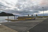 a sidewalk leading to the waterfront of a river with mountains in the background with a road and pier