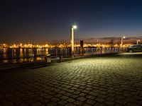 cars parked at night next to the harbor on brick walkways near water and lights