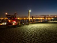 cars parked at night next to the harbor on brick walkways near water and lights