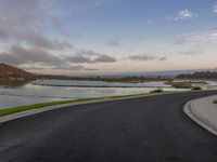 an empty highway next to the shore near water with a hill in the background that has some grass