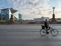 a woman riding a bike across a bridge with tall buildings behind her and the person in the bike is driving