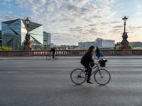 a woman riding a bike across a bridge with tall buildings behind her and the person in the bike is driving