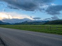 the road is empty during the day and has mountains in the distance as well as a fence