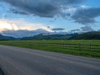 the road is empty during the day and has mountains in the distance as well as a fence