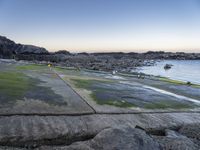 the view is full of people on the water, and the beach has small boats and a grassy area with large rocks