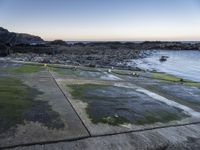 the view is full of people on the water, and the beach has small boats and a grassy area with large rocks