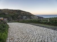 a paved road near the ocean by a rocky hilltop, surrounded by vegetation, with a bench in the distance