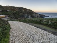 a paved road near the ocean by a rocky hilltop, surrounded by vegetation, with a bench in the distance