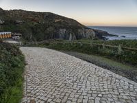 a paved road near the ocean by a rocky hilltop, surrounded by vegetation, with a bench in the distance